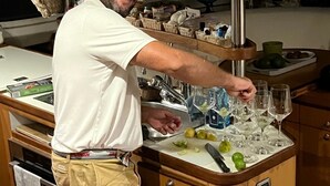 Une personne vêtue d’une chemise blanche et d’un short kaki prépare des boissons dans un bar doté d’un comptoir en bois, d’un évier et de diverses bouteilles et verres.