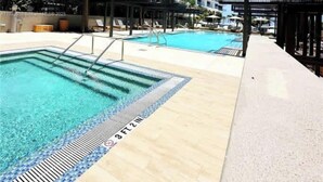 A hotel with a pool area, including a large pool and a smaller pool, surrounded by palm trees and a clear blue sky.