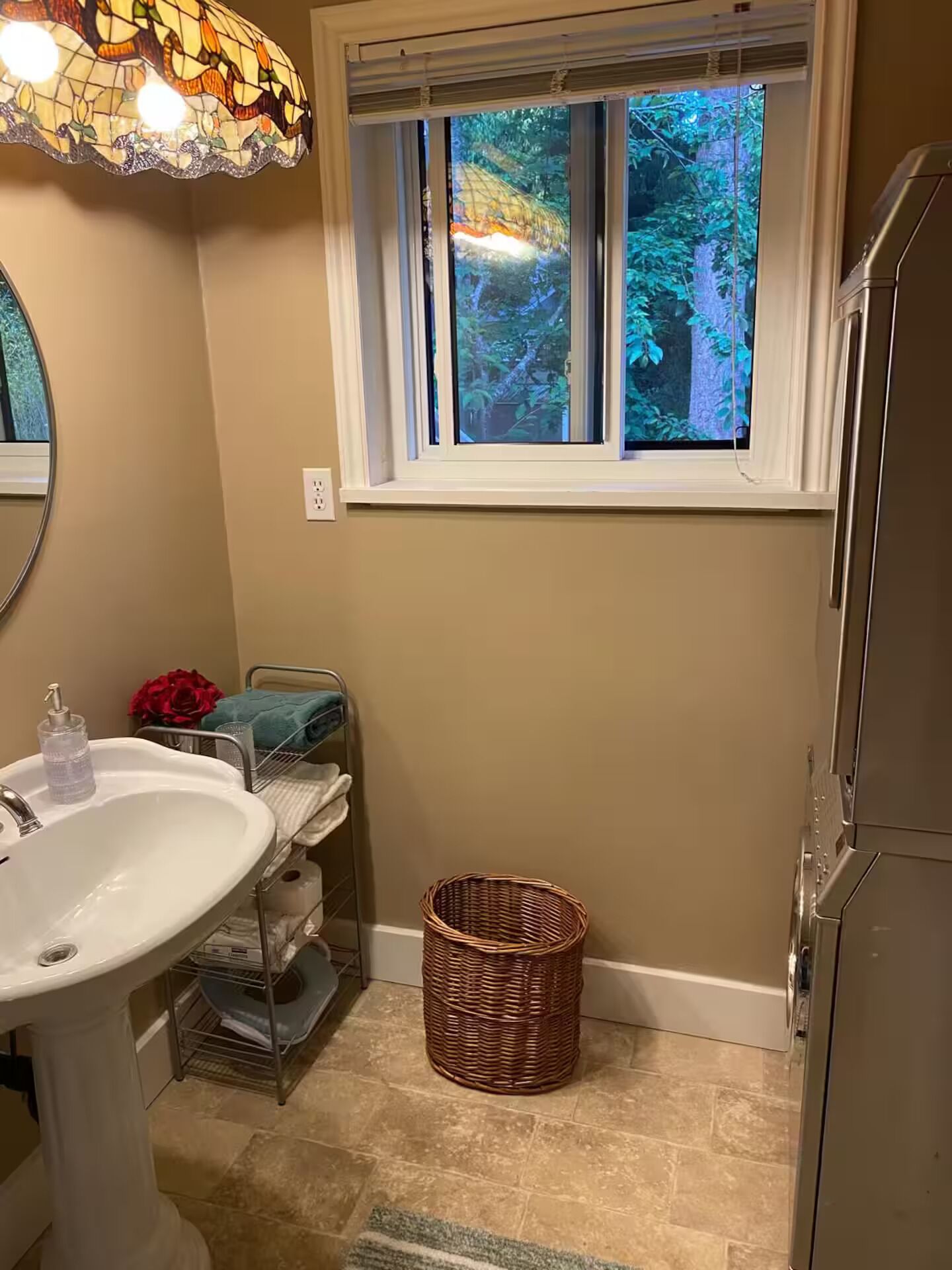 Ein Badezimmer mit weißem Waschbecken, einem Fenster mit Blick auf Grünflächen, einem Korbwäscheschrank und einer Duschkabine.