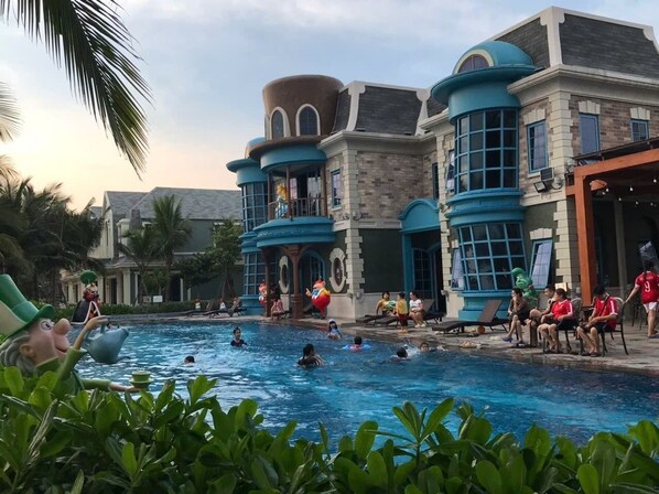 Zona de piscina con gente nadando y relajándose, rodeada de edificios de arquitectura singular.