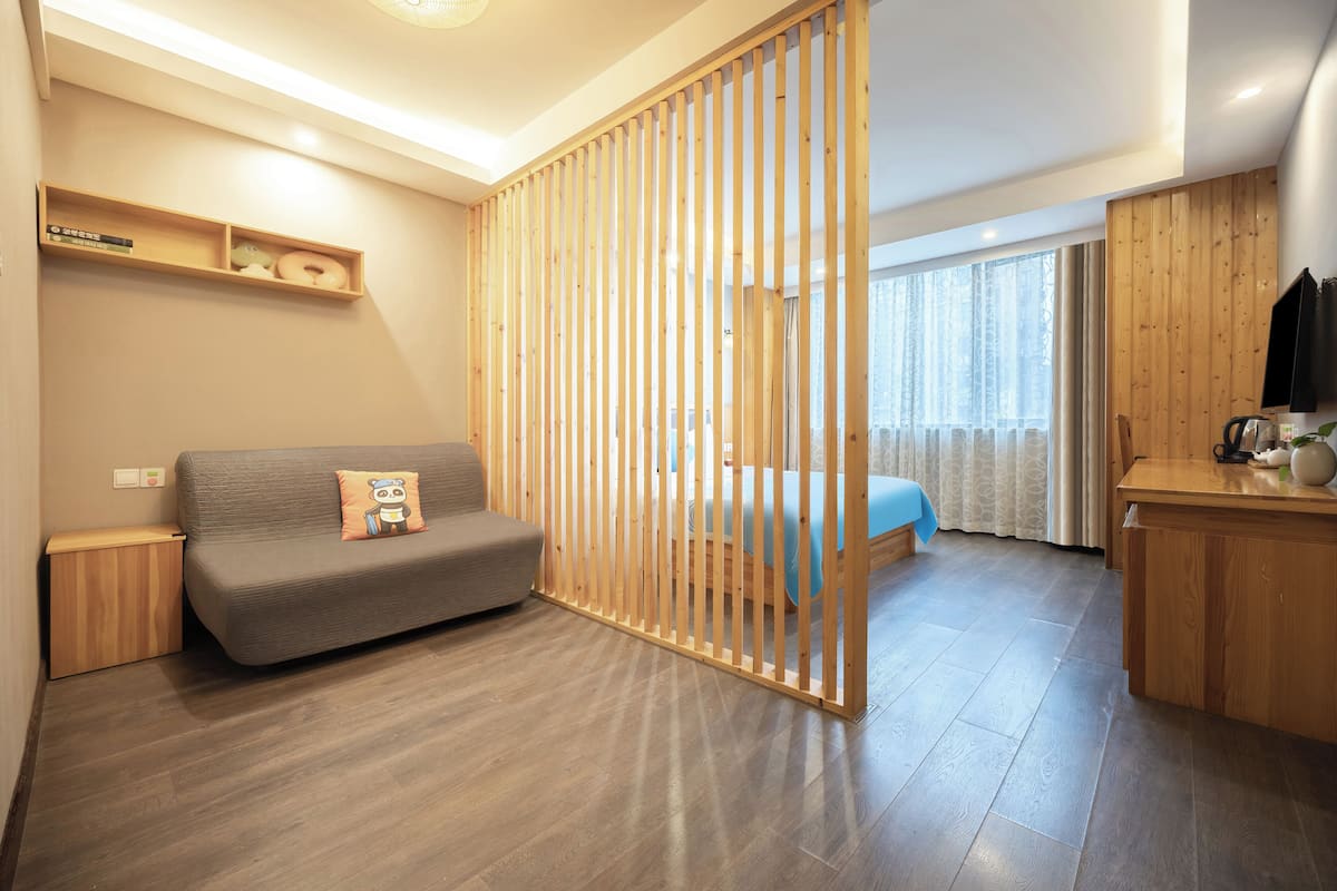 Una habitación moderna con sofá gris, una división de madera, un mueble de madera pequeño, una cama con colcha azul y un escritorio de madera con hervidor.