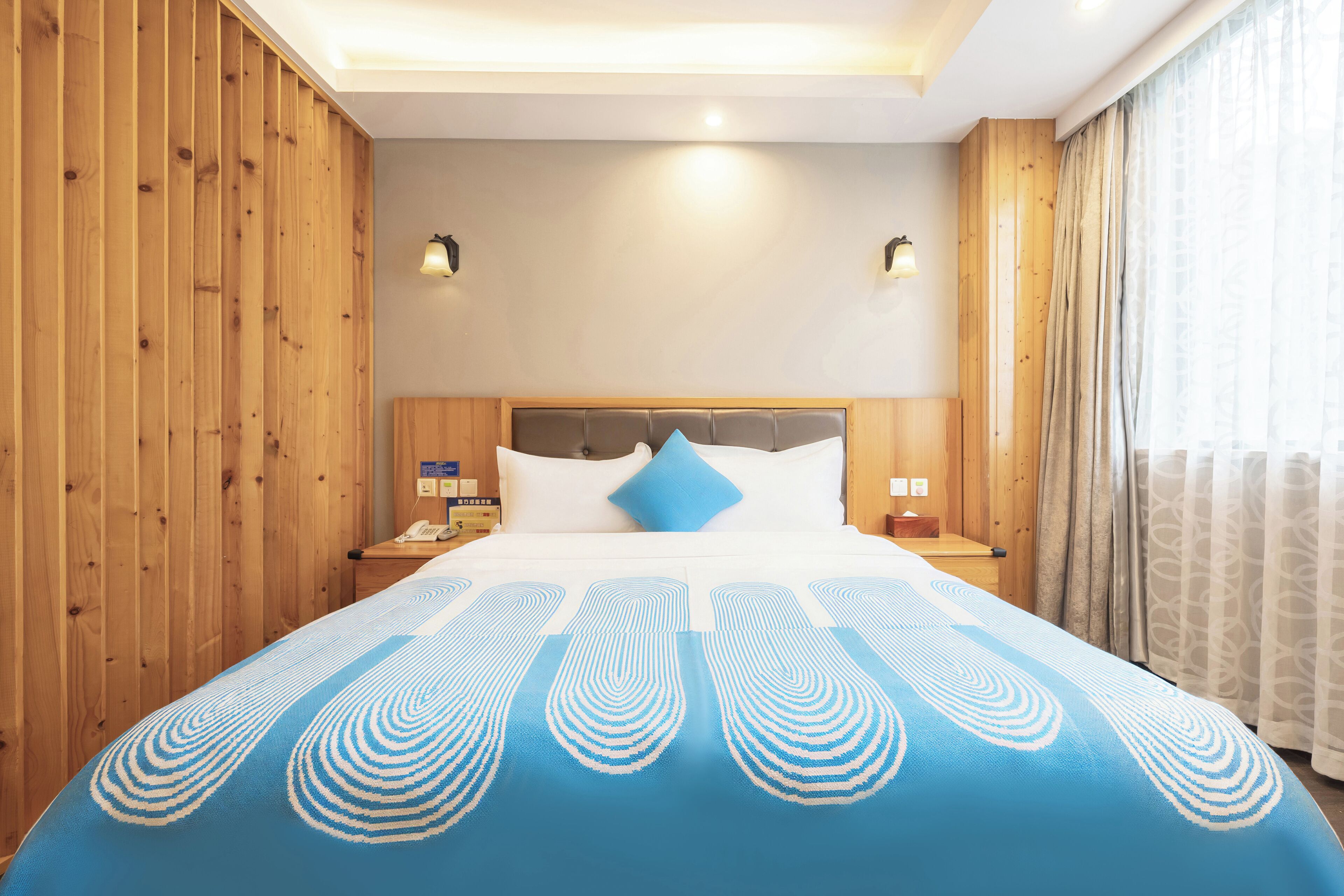 Une chambre d’hôtel avec un mur recouvert de panneaux en bois, un grand lit avec une couvre-lit à motifs bleus et blancs, deux tables de chevet avec des lampes et une fenêtre avec des rideaux.