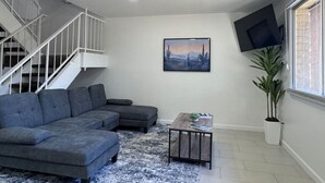Una sala de estar con un sofá modular, una mesa de centro, un ventilador de techo, una escalera, un televisor montado y una planta en maceta.
