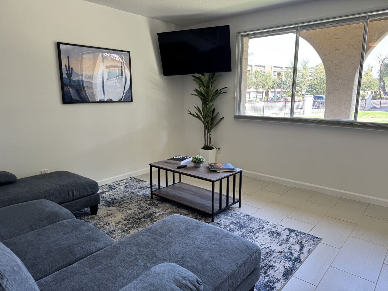 A living room with a sofa set, a coffee table, a TV mounted on the wall, and a window with a view of the outside.