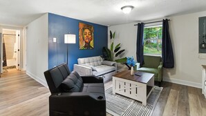 A modern living room with a blue accent wall, a white sofa, a black leather chair, and a green armchair. There is a painting on the wall, a floor lamp, and a window with curtains. - Mountain Bungalow | Nature, History| Wineries|PS5 (Harpers Ferry)