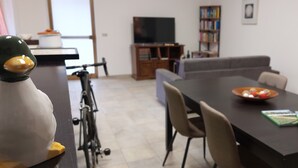 Un coin repas avec une table, des chaises et un saladier rempli de fruits. On aperçoit une bibliothèque et un téléviseur au fond.