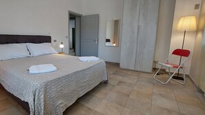 Un lit bien fait, avec une couvre-lit à motifs et des oreillers, une table de chevet avec une lampe, une chaise pliante rouge et un miroir accroché au mur.