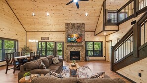 A spacious living room with a stone fireplace, wooden ceiling, and large windows. - Dollywood 28 min | Game Room + Hot Tub + Fire Pit (Sevier County)