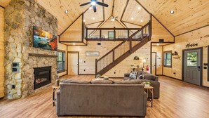 A spacious living area with a stone fireplace, a large sofa, a wooden staircase, and a ceiling fan. - Dollywood 28 min | Game Room + Hot Tub + Fire Pit (Sevier County)