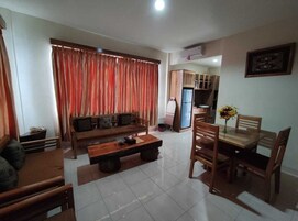 Una sala de estar con sofá, mesa de centro y mesa de comedor con sillas. Se observa una zona de cocina al fondo.