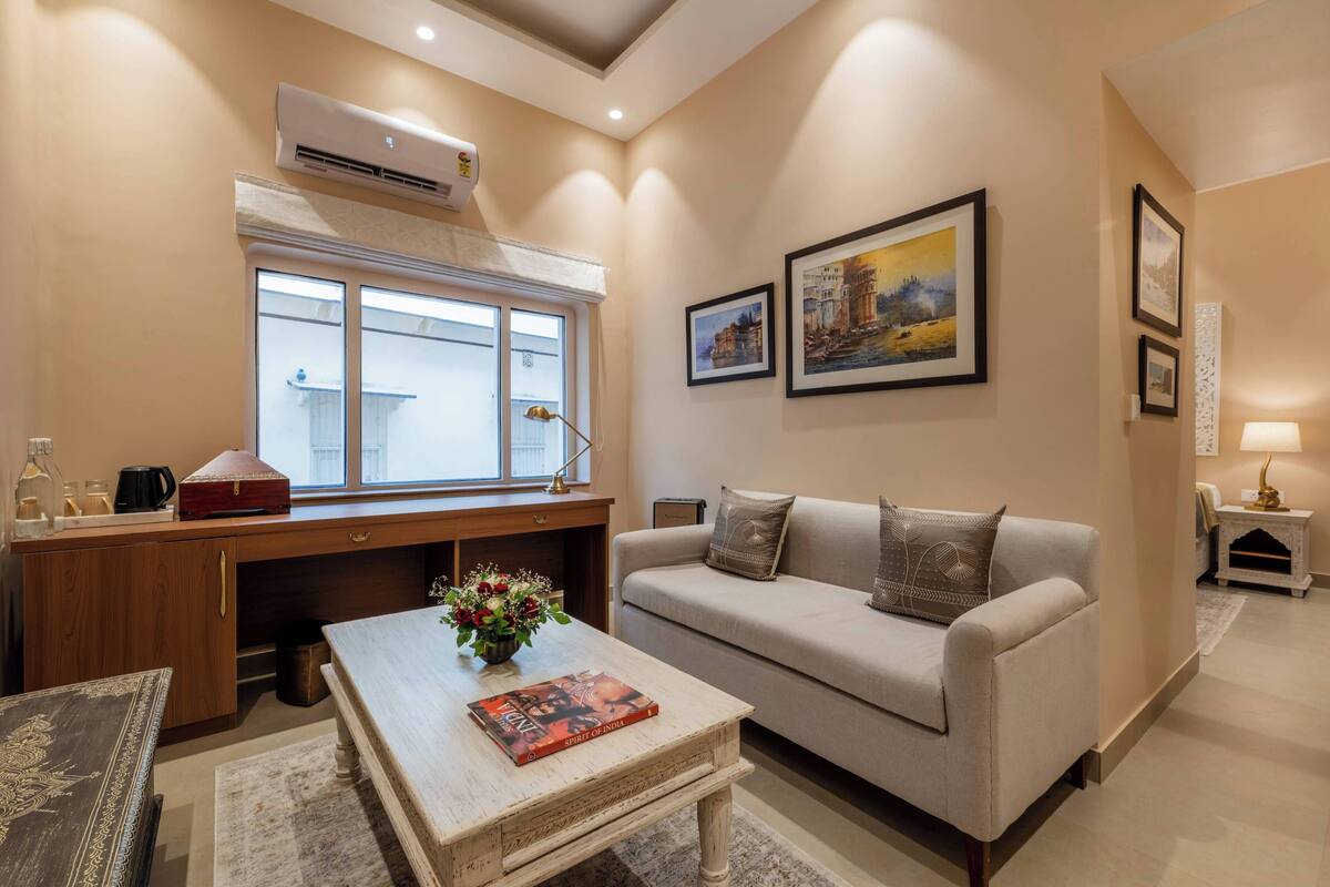 A living room with a sofa, coffee table, and a desk with a computer. There is a window with blinds, a lamp, and framed pictures on the wall.
