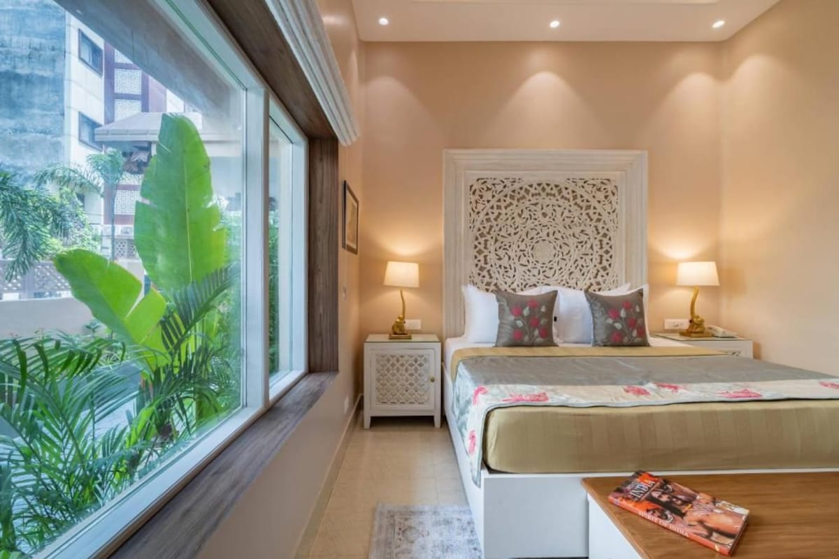 A bedroom with a bed, two bedside lamps, a nightstand, a window with a view of greenery, and a magazine on the bed.