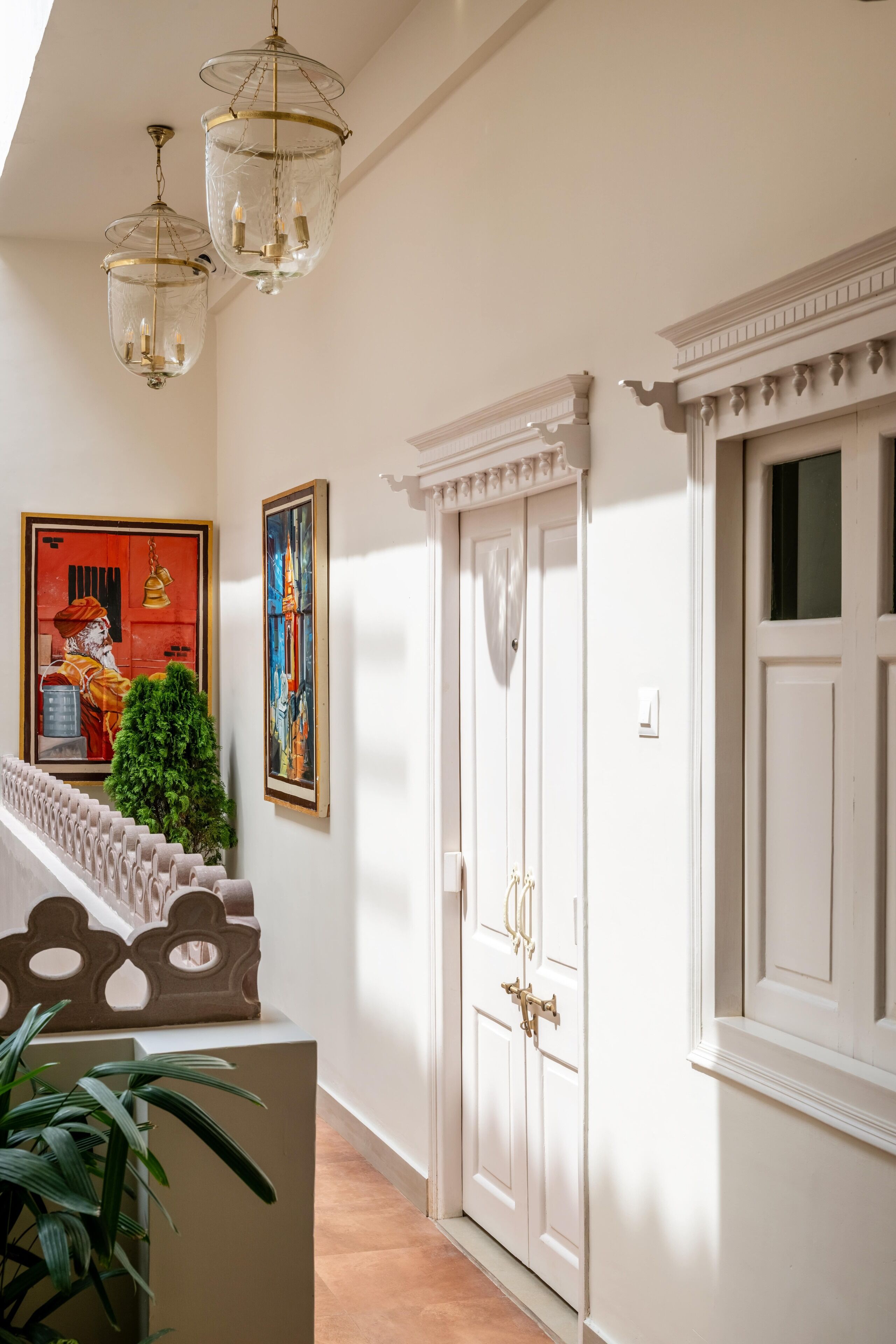 A hallway with white walls, a staircase with a decorative railing, and two chandeliers. There are paintings on the wall and a door with a decorative trim.