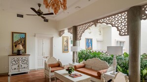 A spacious room with a white sofa set, a coffee table, and a decorative cabinet. There is a ceiling fan, a chandelier, and a view of greenery outside. - juSTa Luxe - Kashi Parampara, Varanasi (Varanasi)