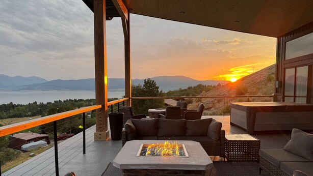 A modern outdoor living area with a stone fireplace, comfortable seating, and a view of a sunset over a lake. - NEW in 2026! Elegant, Dramatic Mountain/Lake Views! 20 Mins to Yellowstone NP! (Island Park)