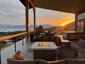 A modern outdoor living area with a stone fireplace, comfortable seating, and a view of a sunset over a lake. - NEW in 2026! Elegant, Dramatic Mountain/Lake Views! 20 Mins to Yellowstone NP! (Island Park)