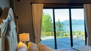 A bedroom with a large bed, a bedside lamp, and a view of a balcony with outdoor furniture. - NEW in 2026! Elegant, Dramatic Mountain/Lake Views! 20 Mins to Yellowstone NP! (Island Park)