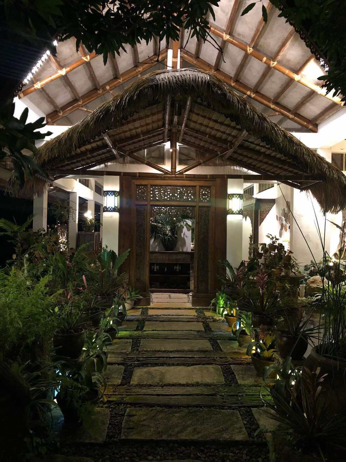 A covered walkway with a thatched roof, surrounded by potted plants and a stone pathway.