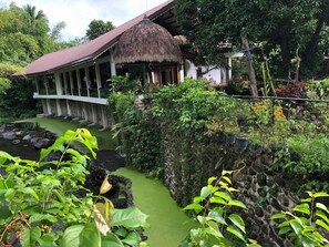 A traditional thatched-roof structure with a white exterior, surrounded by lush greenery and a small stream. - Harmonia Nature Resort by Hiverooms (Majayjay)