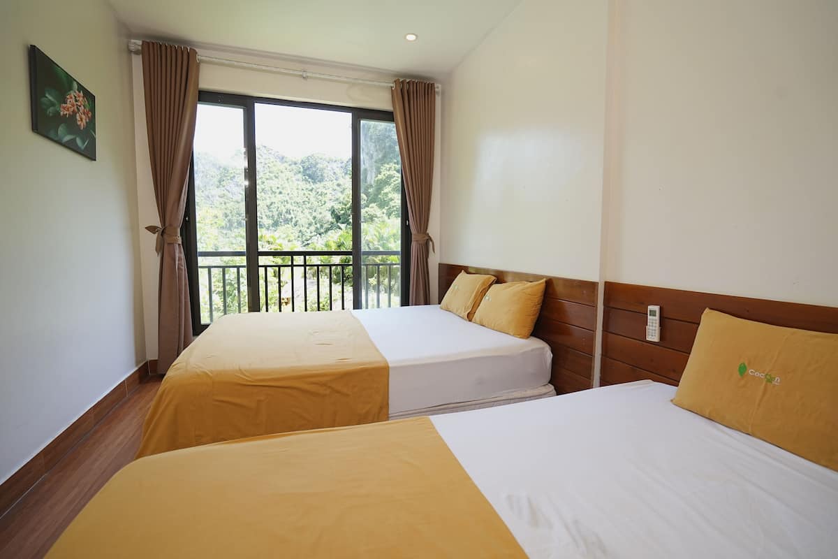 A hotel room with two beds, a balcony with a view of greenery, and a painting on the wall.