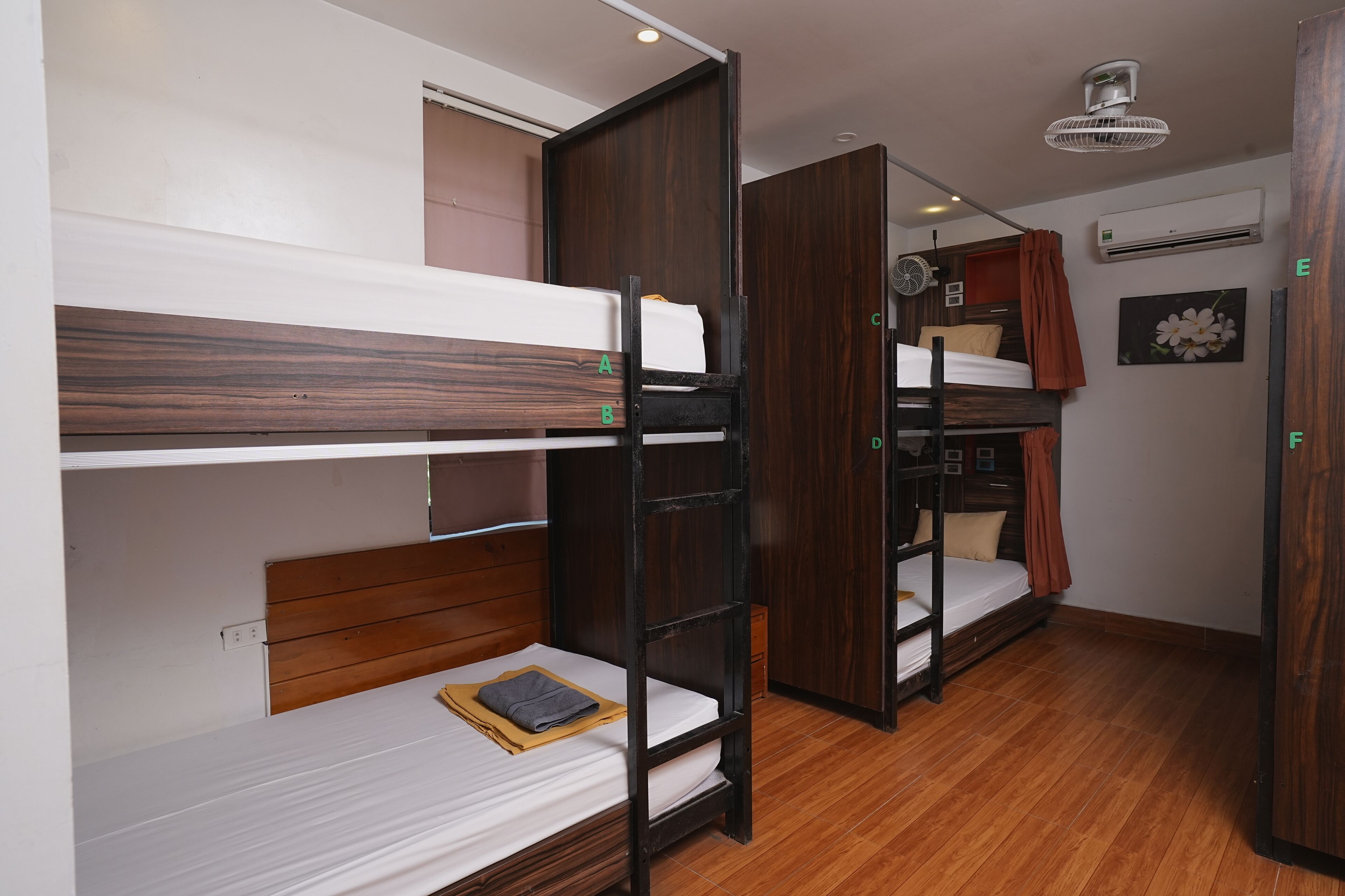 A dormitory room with bunk beds, wooden flooring, a ceiling fan, and an air conditioning unit.