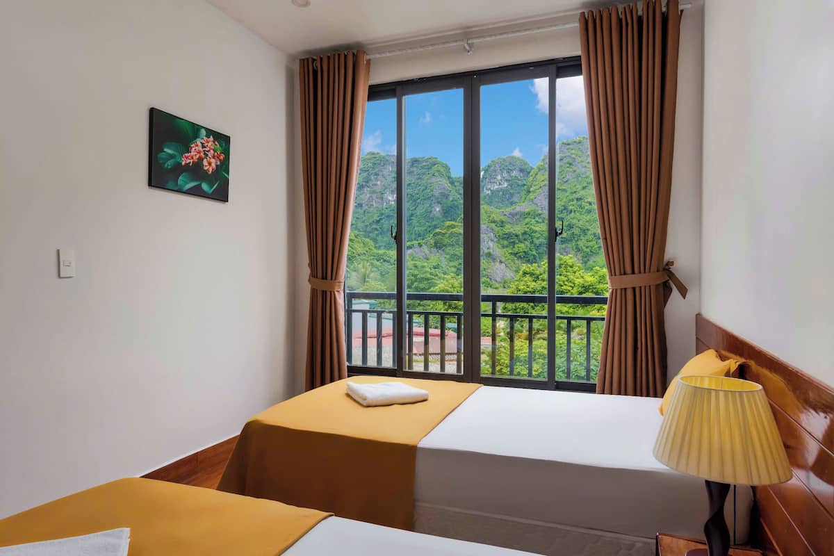 A hotel room with two beds, a window with curtains, a view of green mountains, and a lamp on the nightstand.