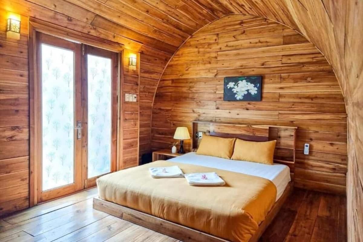 A cozy wooden cabin room with a bed, a nightstand, and a window with a view of trees.