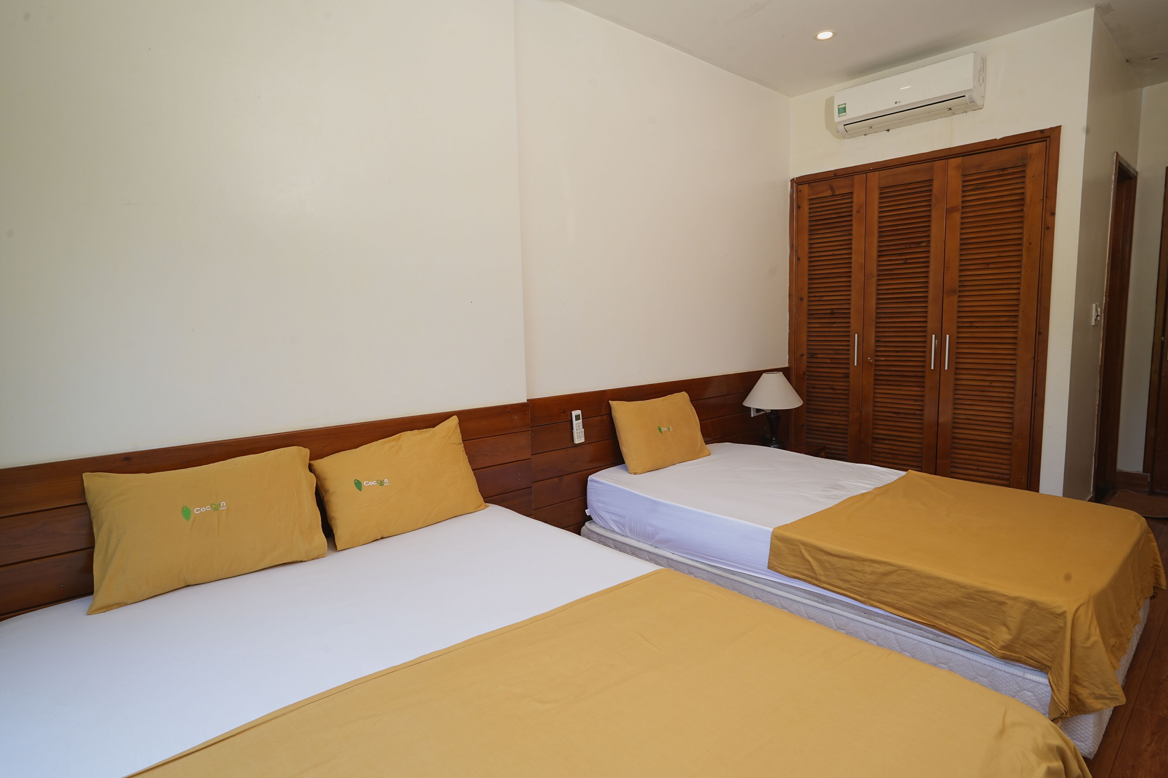 A hotel room with two beds, each with a white mattress and a brown bedspread. There is a wooden wardrobe and an air conditioning unit on the wall.