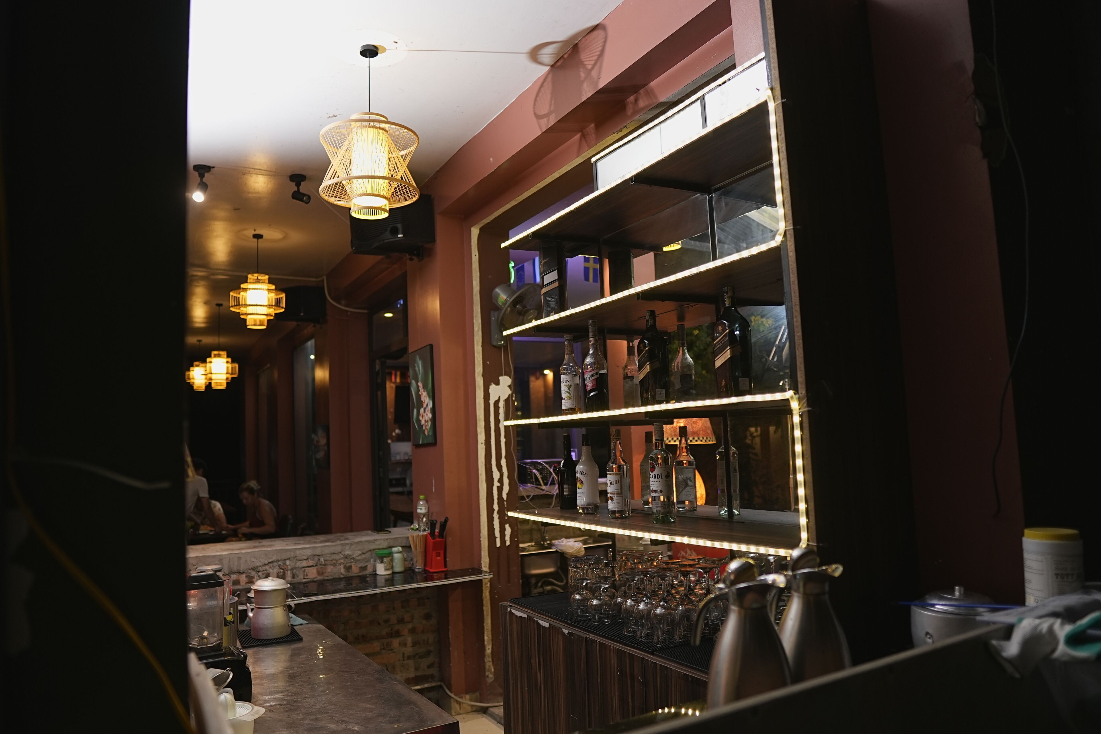 A bar area with shelves of bottles, a counter with glasses, and hanging lights.