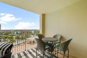 A balcony with wicker chairs and a small table, overlooking a pool and buildings.