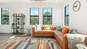 A modern living room with a sectional sofa, a colorful rug, a bookshelf, and large windows.