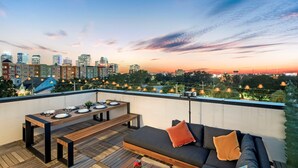 A rooftop terrace with a dining table, benches, and a sofa, overlooking a city skyline at sunset.