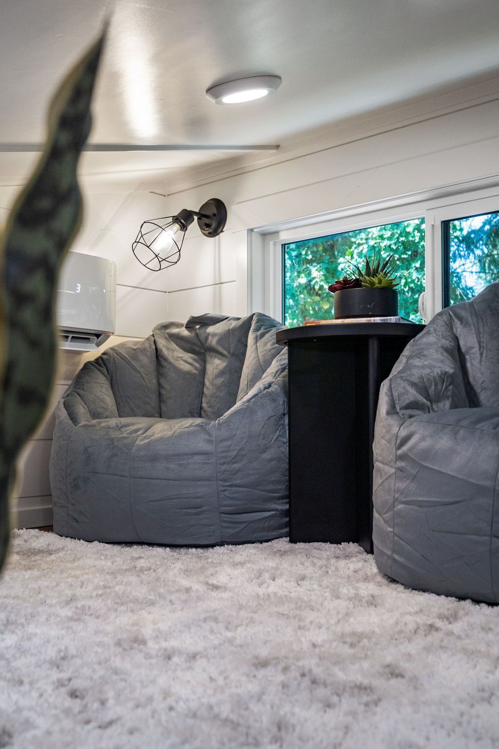 A modern living room with a white shag carpet, a grey bean bag chair, a black side table with a potted plant, and a window with a view of greenery.