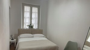 Ein Schlafzimmer mit Bett, Nachttisch, Sessel, Fenster mit Vorhängen und einer Zimmerpflanze.