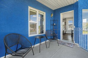 Un mur extérieur bleu avec une porte blanche, une chaise en métal noir, une petite table et une étagère en métal.