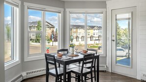 Un comedor con una mesa dispuesta para cuatro personas, ventanas amplias y vistas a un barrio residencial.