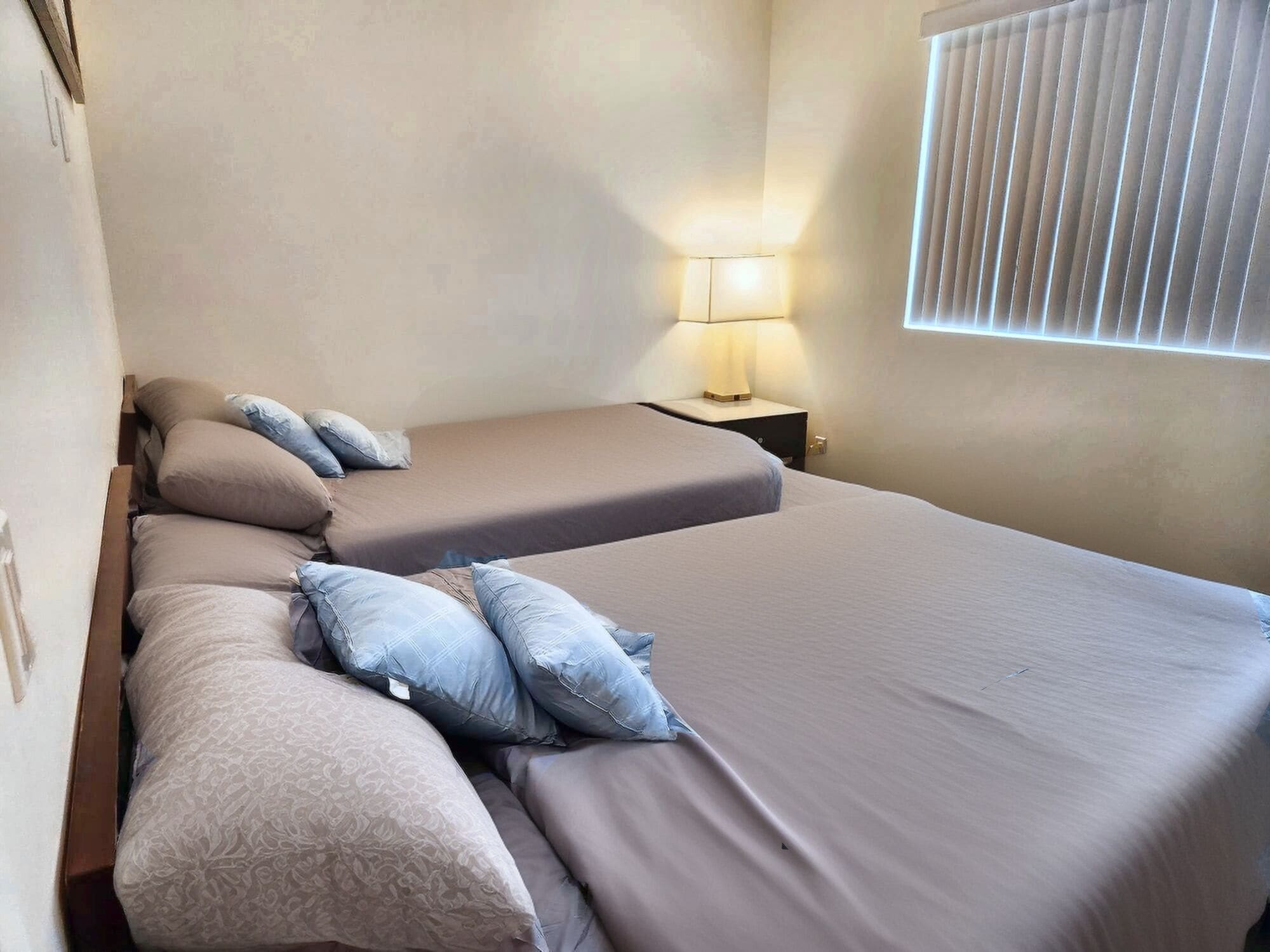 A hotel room with two single beds, a nightstand with a lamp, and a window with blinds.