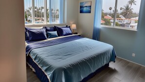 Un dormitorio con una cama grande, dos ventanas con cortinas azules, una mesita de noche con una lámpara y un cuadro en la pared.
