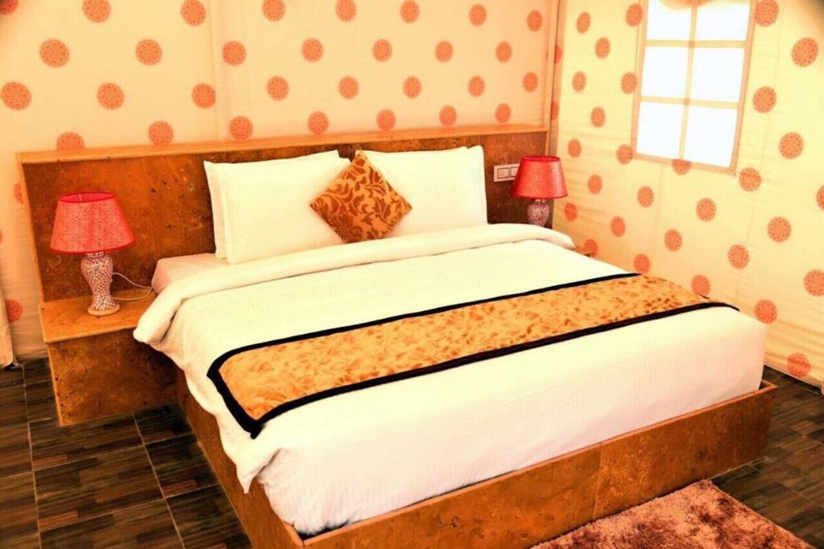Une chambre avec un lit en bois, du linge de lit blanc, un plaid à motifs, une lampe rouge et un papier peint à pois.