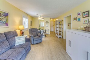 A living room with a sofa, armchair, and a lamp. There is a dining table with chairs and a kitchen area with cabinets and a refrigerator.