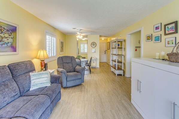 A living room with a sofa, armchair, and a lamp. There is a dining table with chairs and a kitchen area with cabinets and a refrigerator.