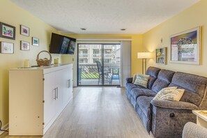 A compact living space with a sofa, a flat-screen TV, a dining table, and a balcony with a view of buildings.
