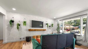 Un salon moderne avec un canapé vert, un téléviseur à écran plat fixé au mur et plusieurs plantes suspendues.