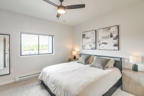 Une chambre à coucher comprenant un lit, deux lampes fixées au mur, un ventilateur de plafond, une grande fenêtre et deux tableaux abstraits.