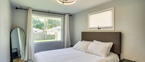 Un dormitorio ordenado con una cama grande, una mesita de noche, un ventilador de techo y una ventana con cortinas.