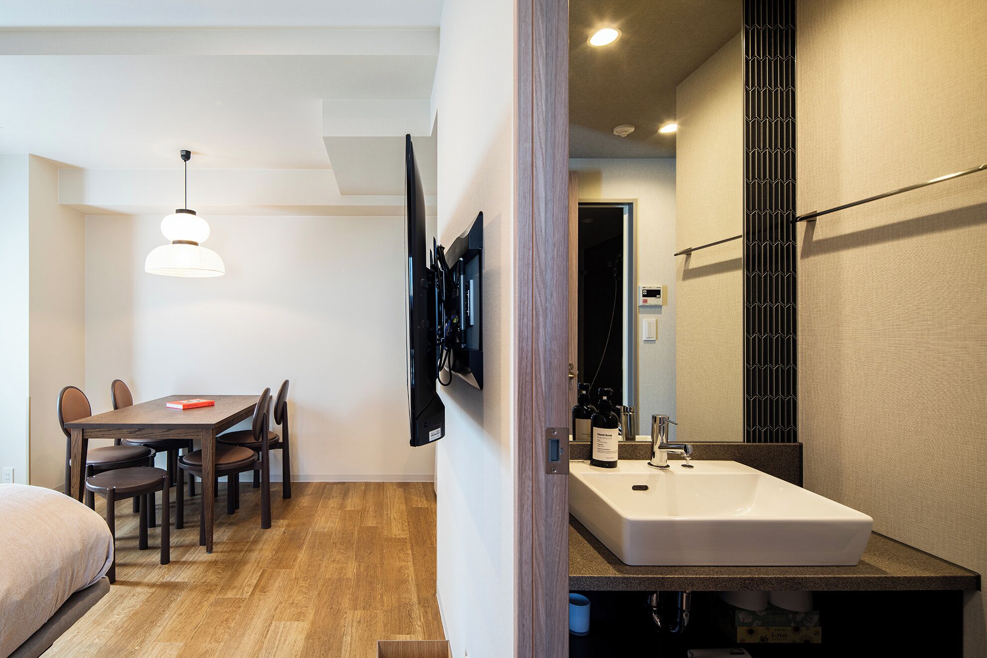 A modern hotel room with a wooden floor, a dining area with a table and chairs, a large mirror, a white sink, and a television mounted on the wall.
