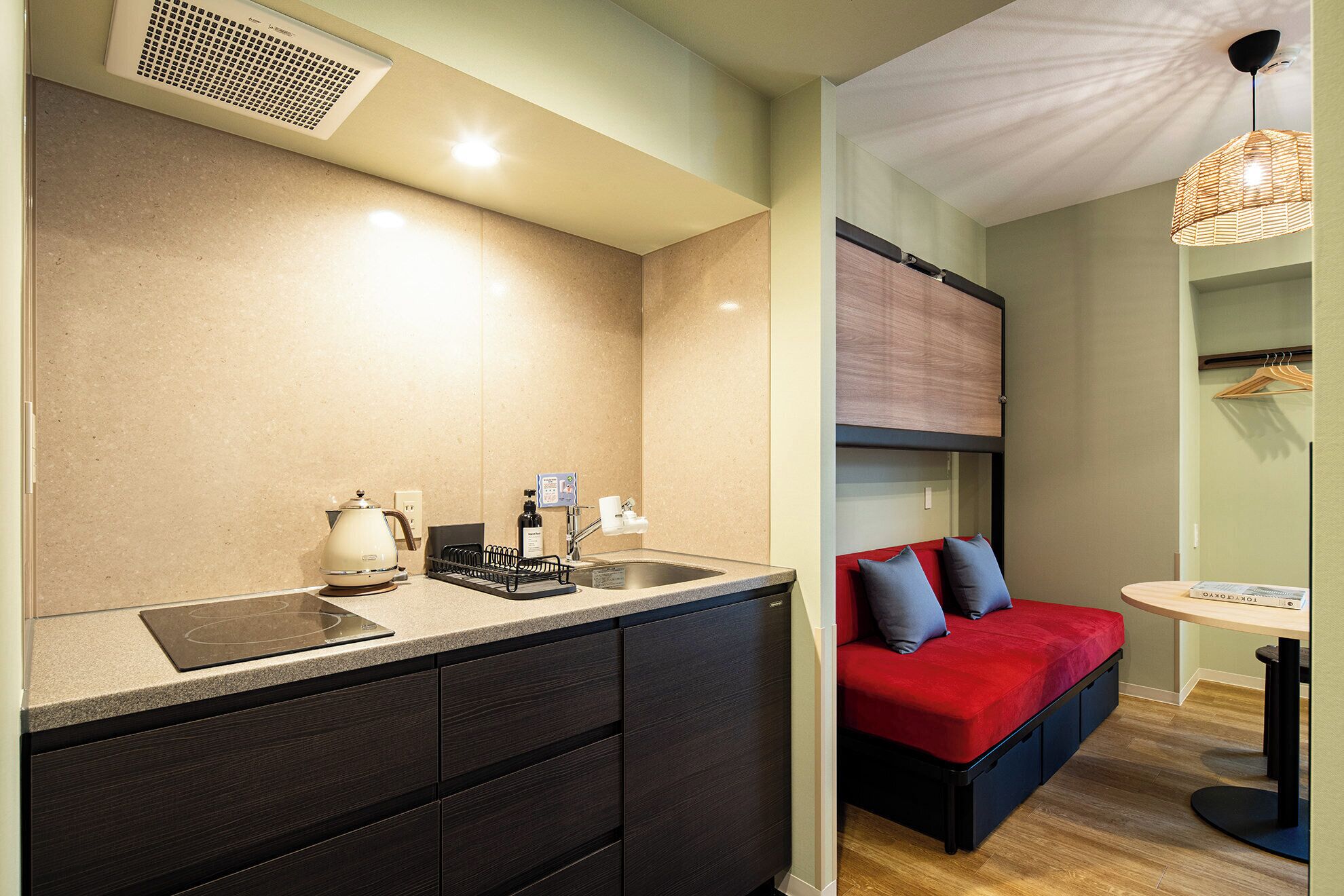 A compact kitchenette with a kettle, coffee maker, and a small sink. There is a red sofa with cushions and a small round table with a magazine.