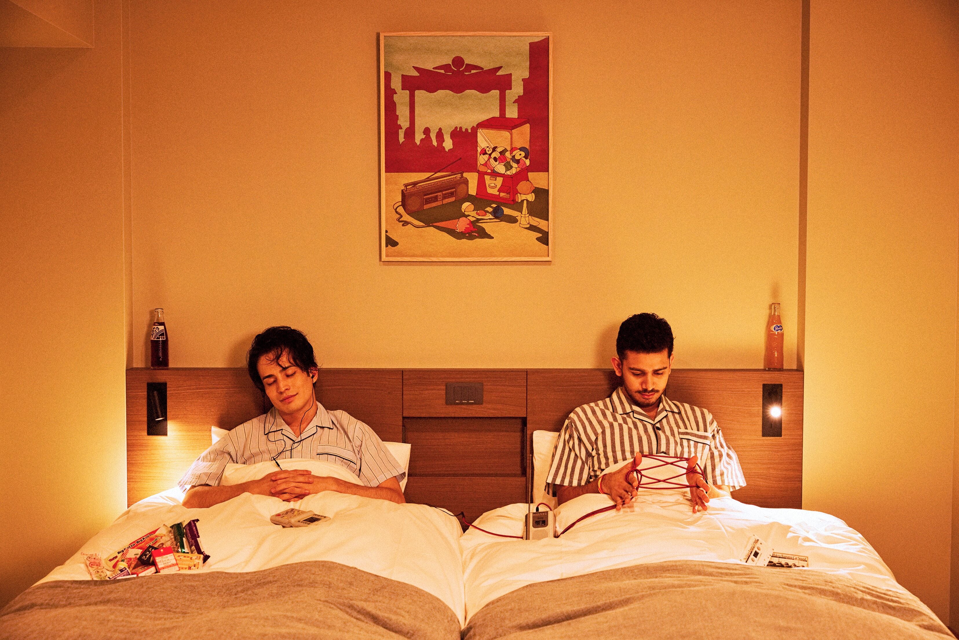 Two men lying in bed in a hotel room, one reading a book and the other using a laptop.