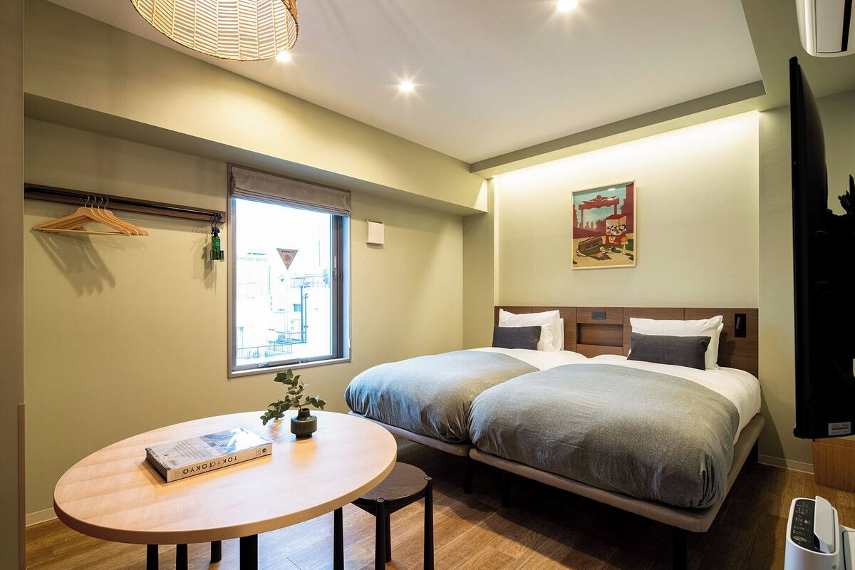 Habitación de hotel con dos camas, una mesa redonda con un libro, una ventana con vistas a edificios y un cuadro en la pared.