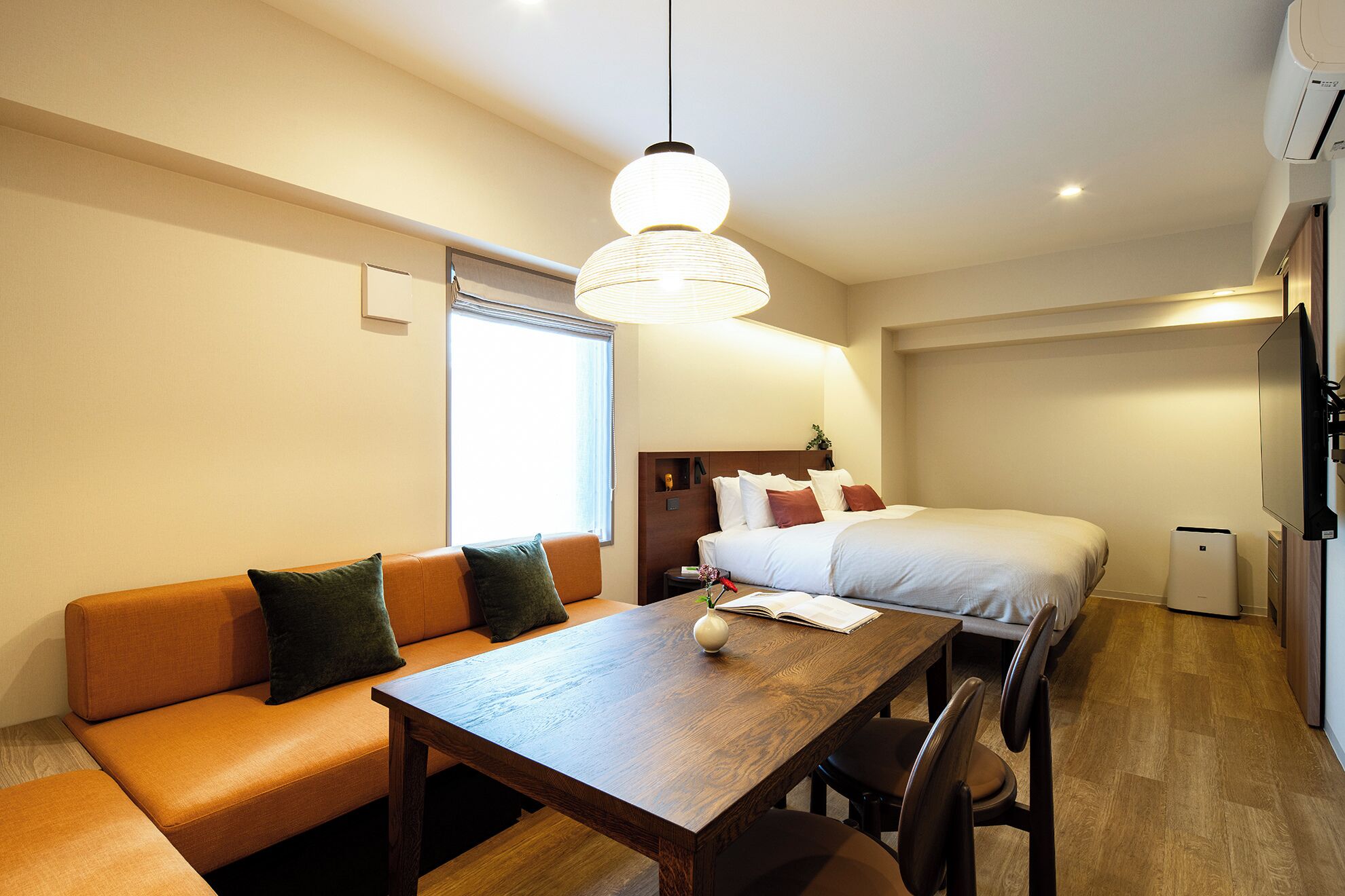 A modern hotel room with a wooden dining table, a sofa, a bed, and a large window.